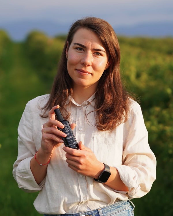 A woman stands in a field, smiling as she holds a Rose Hydration Mist bottle, enjoying the fresh air and nature around her.