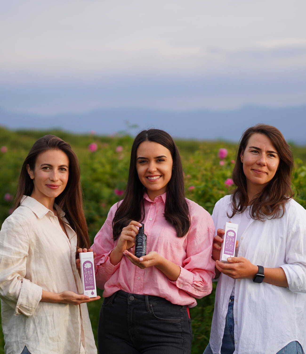 Three women joyfully raising Rose Hydration Mist bottles in front of a picturesque field, celebrating friendship and good times together.