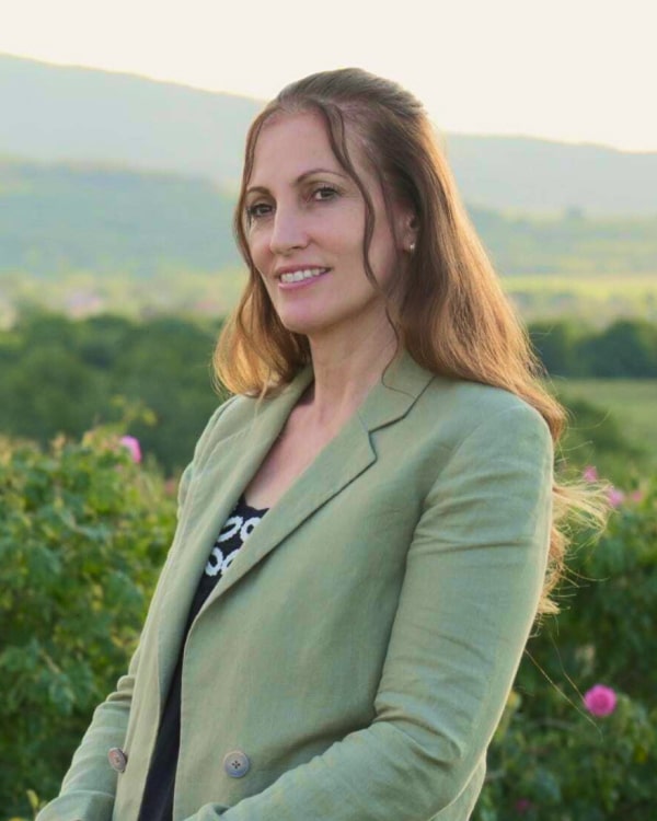 A woman in a green blazer stands confidently in front of a lush green field, enjoying the beautiful outdoor scenery