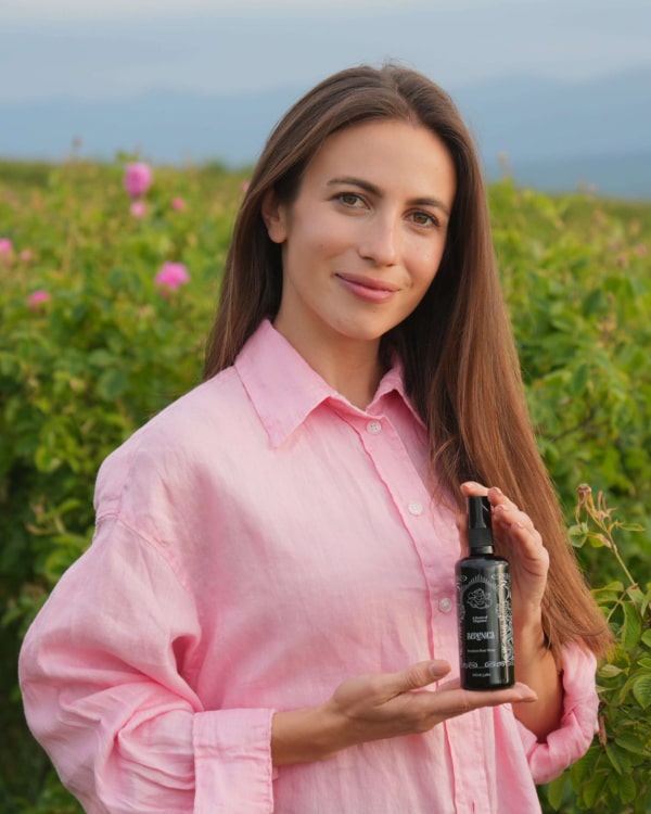 A woman joyfully holds a Rose Hydration Mist bottle while standing in a sunny, green field, surrounded by nature's beauty.